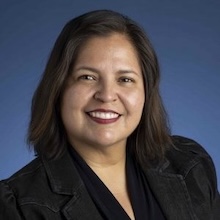 Woman with shoulder length brown hair wearing black blazer smiling blue background